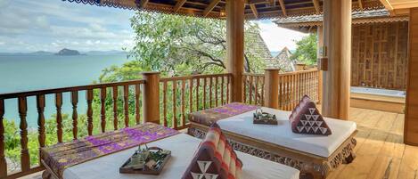 A wooden deck with a view of the sea, furnished with two beds and a table, and a railing.