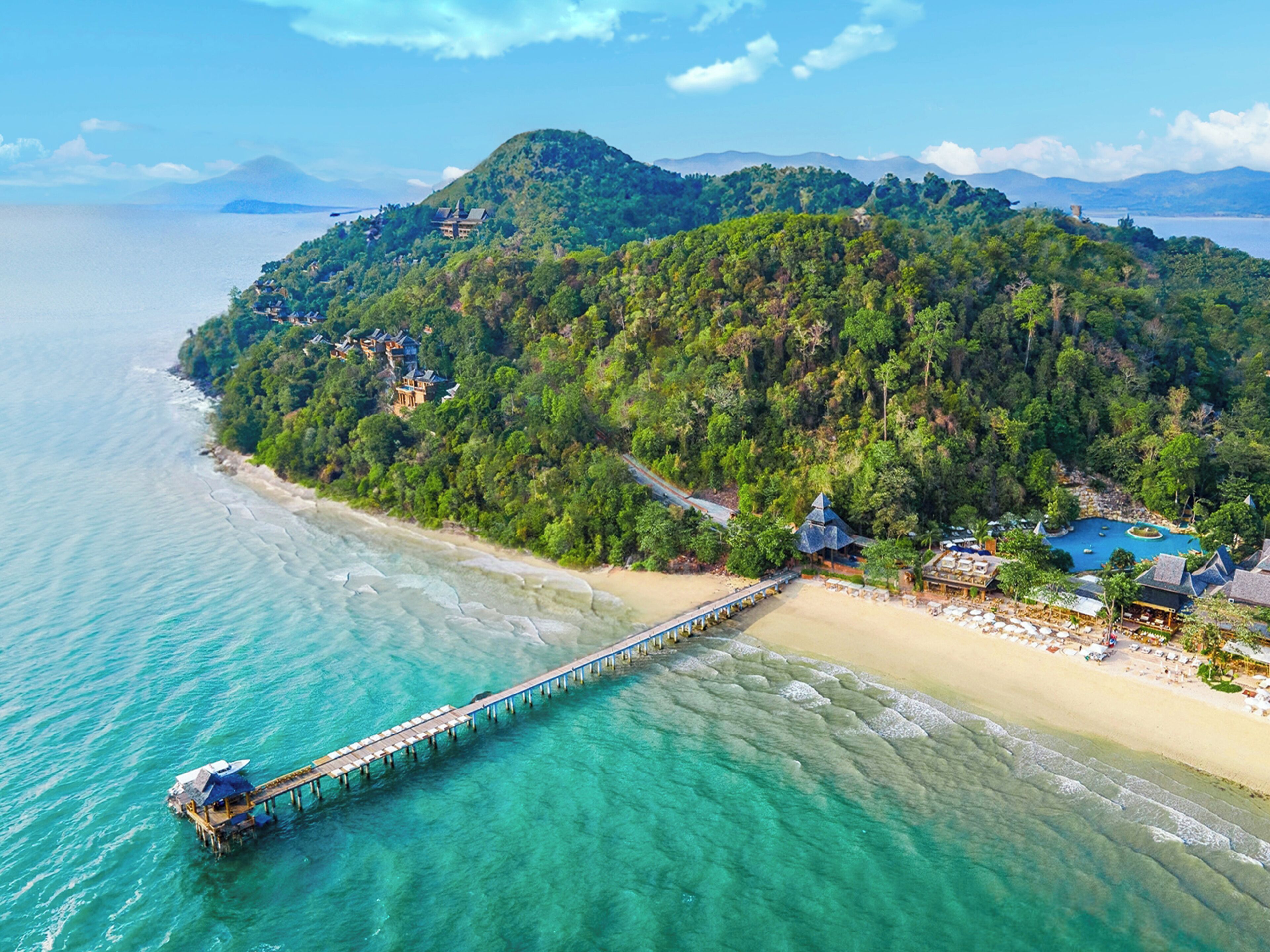 An aerial view of a coastal area with a wooden pier extending into turquoise waters, surrounded by lush greenery and a beach with a resort.