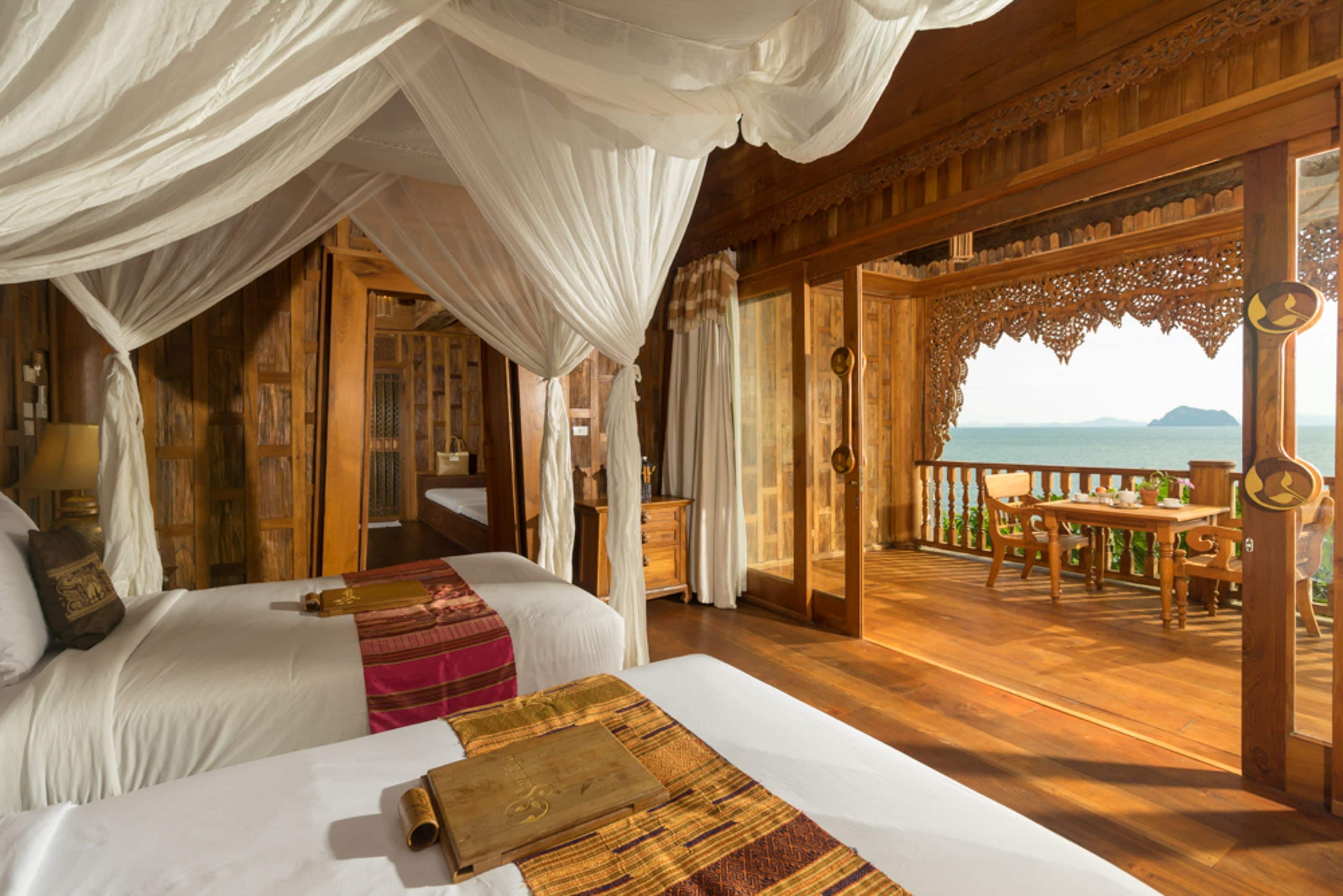 A room with two beds, a balcony with wooden flooring, and a view of the sea.