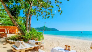 A beachside lounge area with sun loungers and tables, surrounded by greenery and overlooking a clear blue sea.