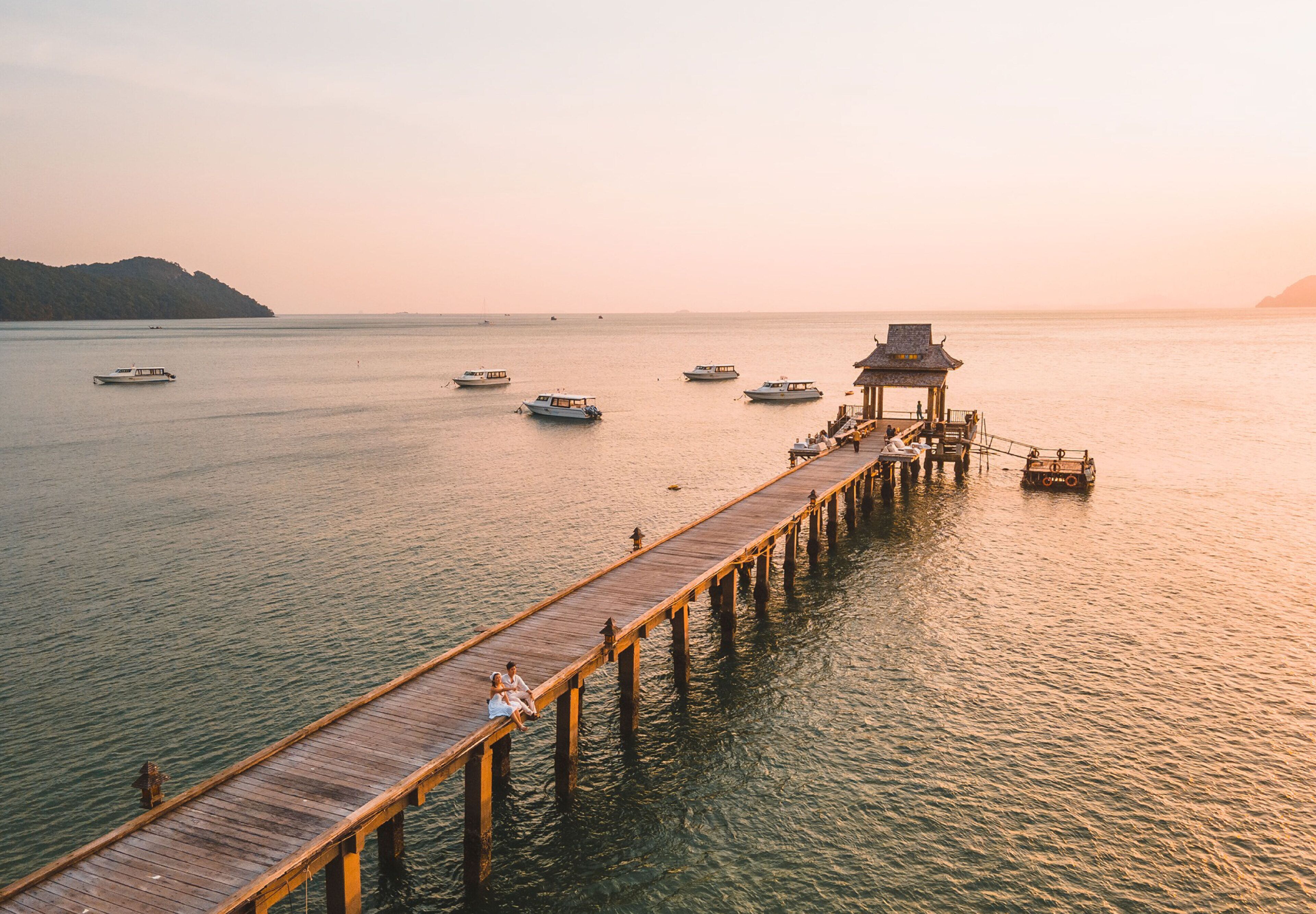 A wooden pier extending into calm waters with boats and a traditional structure at the end.