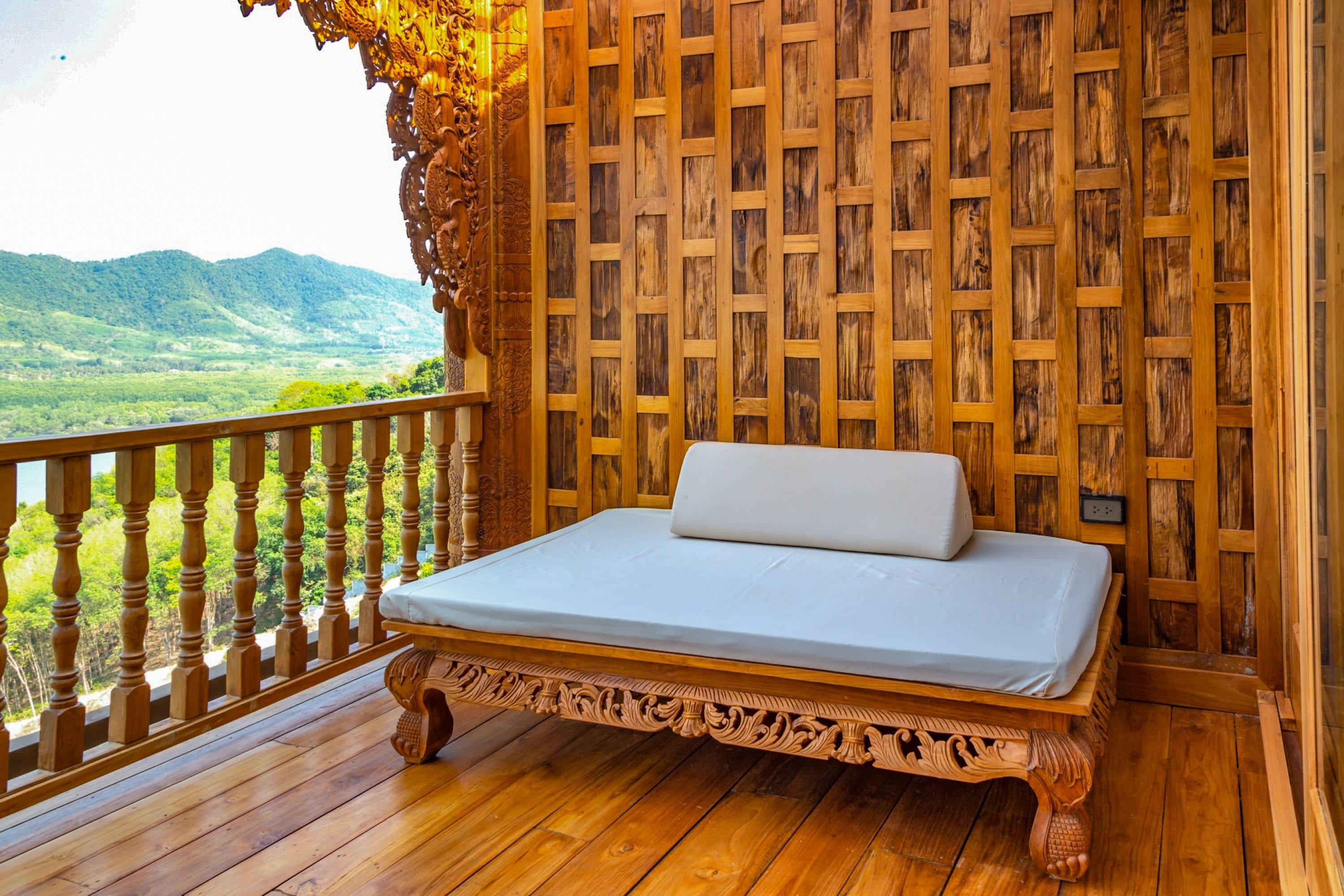 Una cama de madera con colchón blanco en un balcón con barandilla de madera, con vistas a un paisaje montañoso.