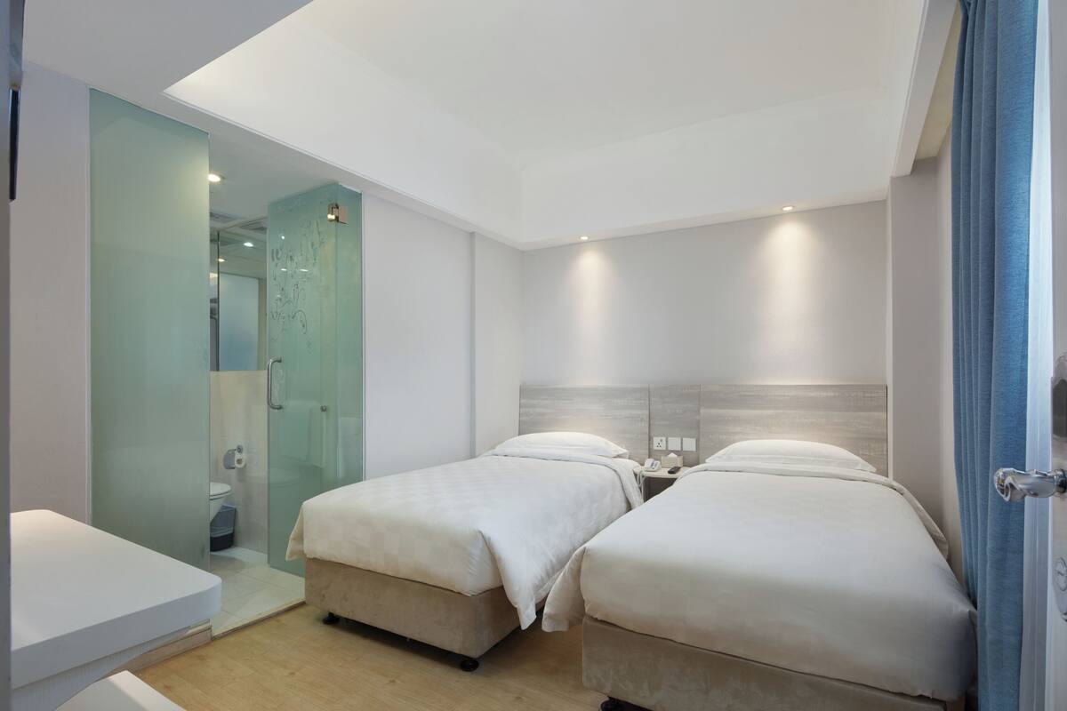 A modern hotel room with two beds, a glass shower enclosure, a small table, and a bedside table with a phone and lamp.