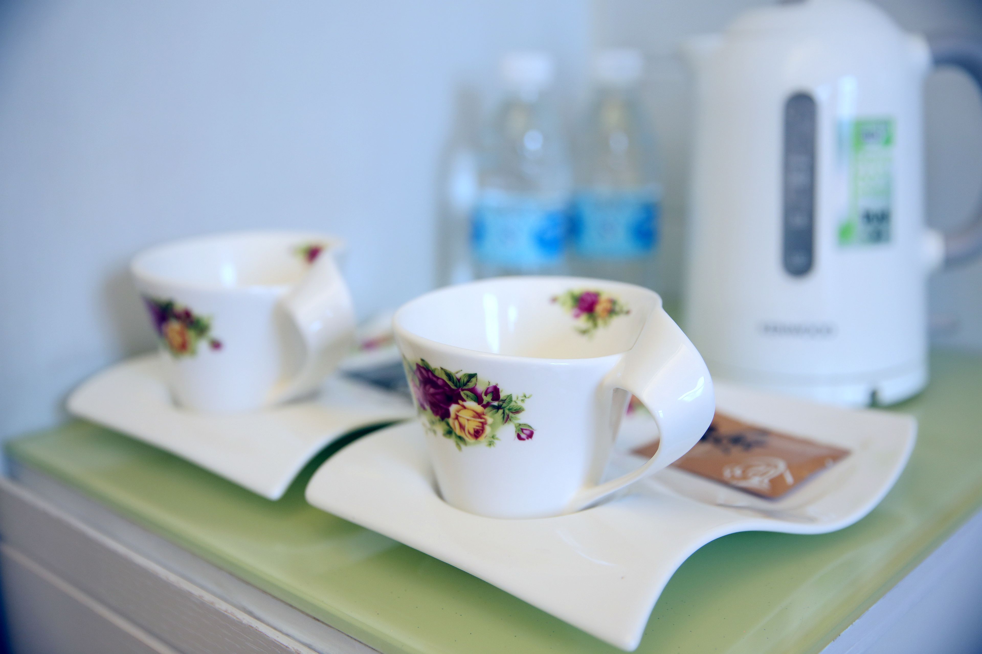 Two white cups with floral designs on a green tray, a white kettle, and a bottle of water in the background.