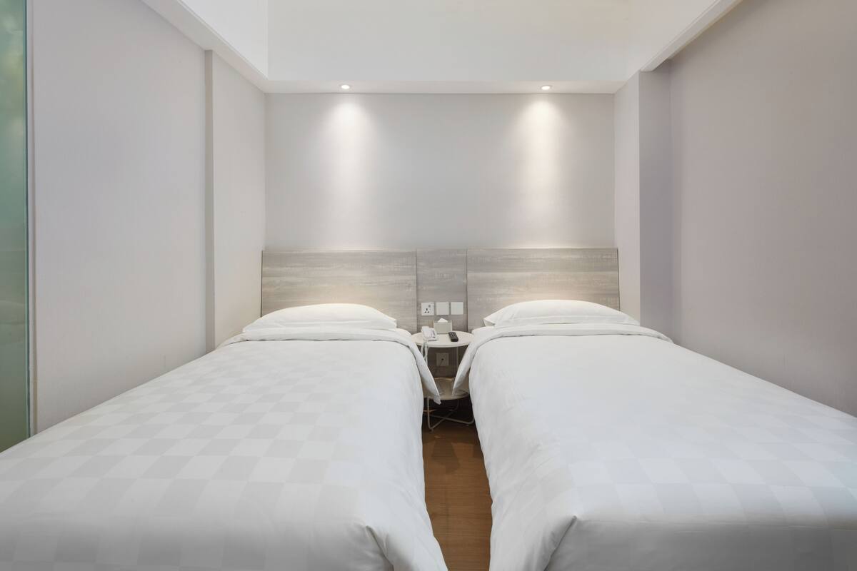 A hotel room with two single beds, a wooden headboard, a bedside table with a lamp, and a small window.