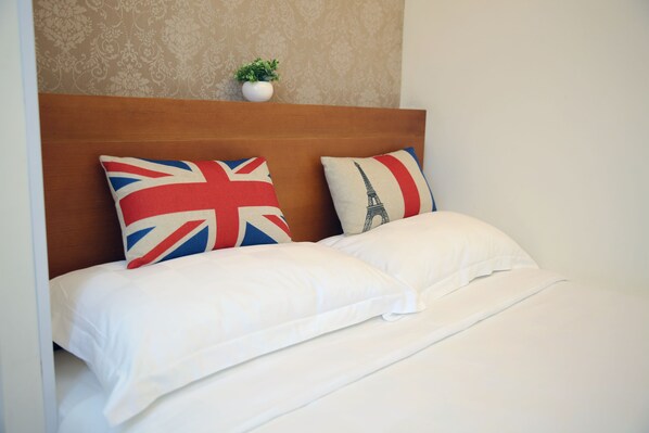 Una cama bien hecha con sábanas blancas y dos almohadas, una con un diseño de la bandera Union Jack y la otra con la Torre Eiffel, en una habitación con cabecera de madera y una pequeña planta en la pared.