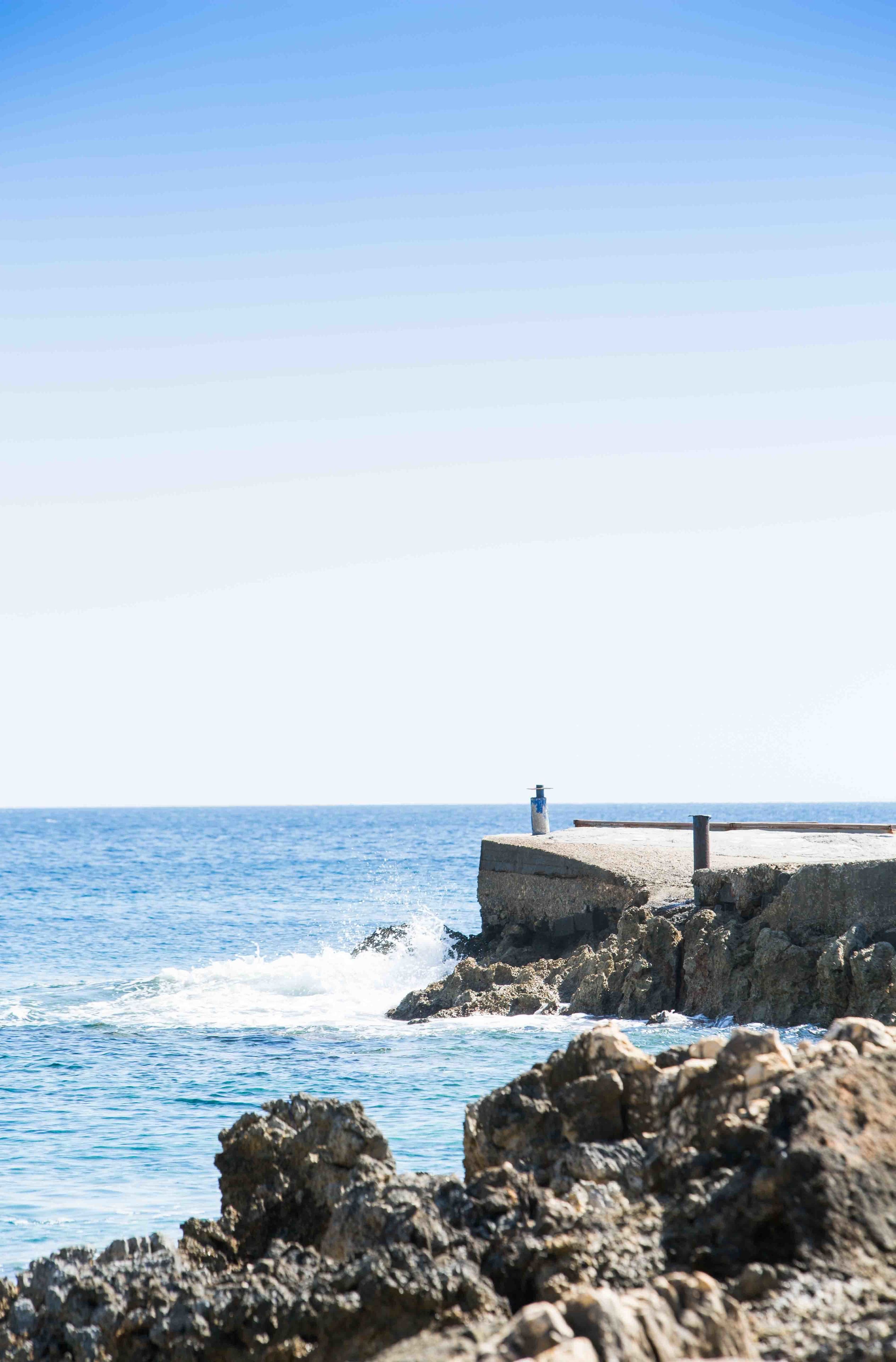 A coastal scene with a concrete pier extending into the sea, surrounded by rocky terrain.