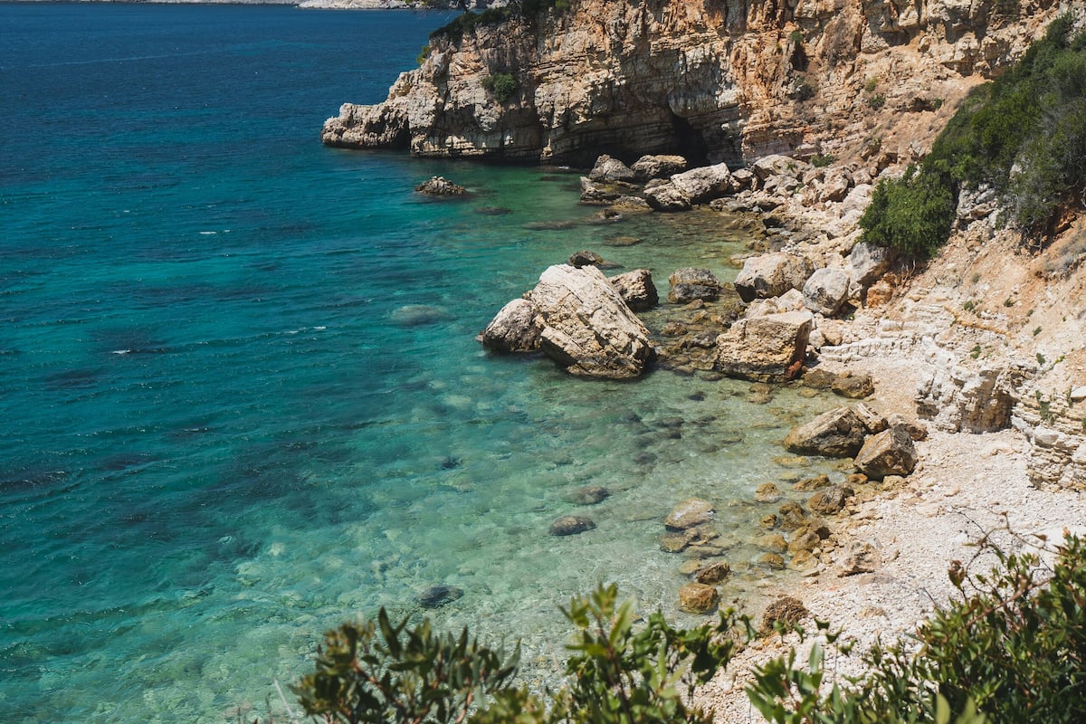 A coastal landscape with clear blue sea, rocky cliffs, and sparse vegetation.