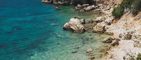 A coastal landscape with clear blue sea, rocky cliffs, and sparse vegetation.
