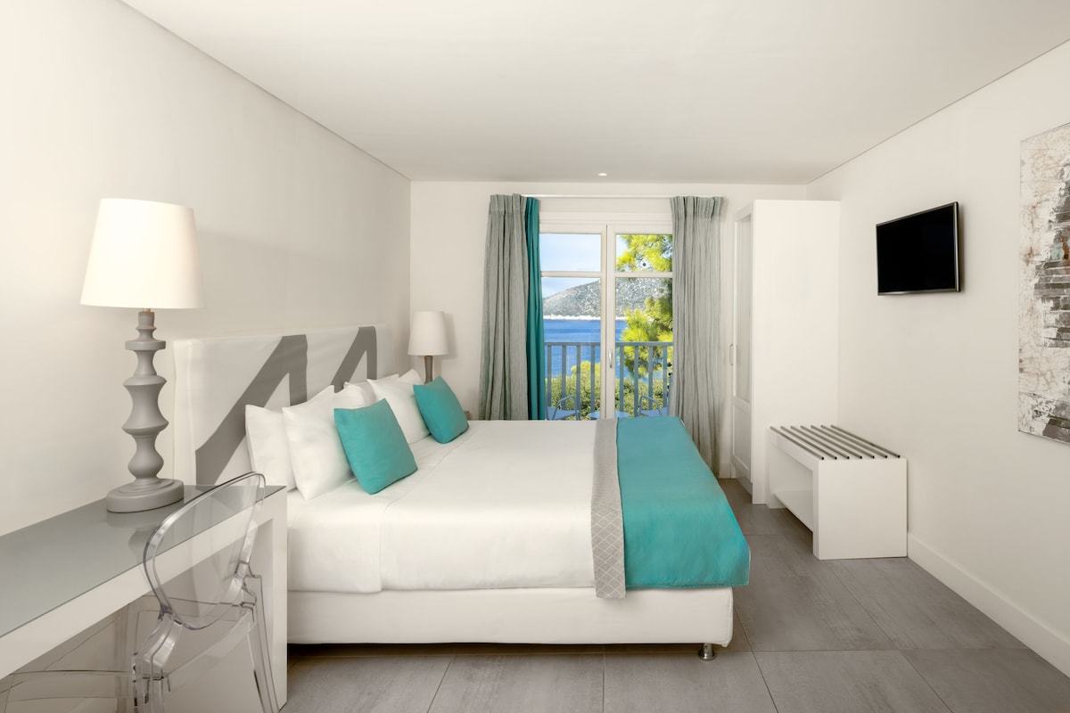 Un dormitorio con cama, televisor, ventana con cortinas y vista al mar.
