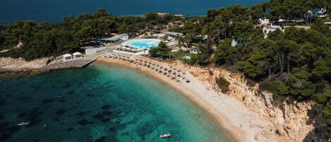 An aerial view of a coastal area with a beach, clear turquoise waters, and a pool area surrounded by trees and buildings.