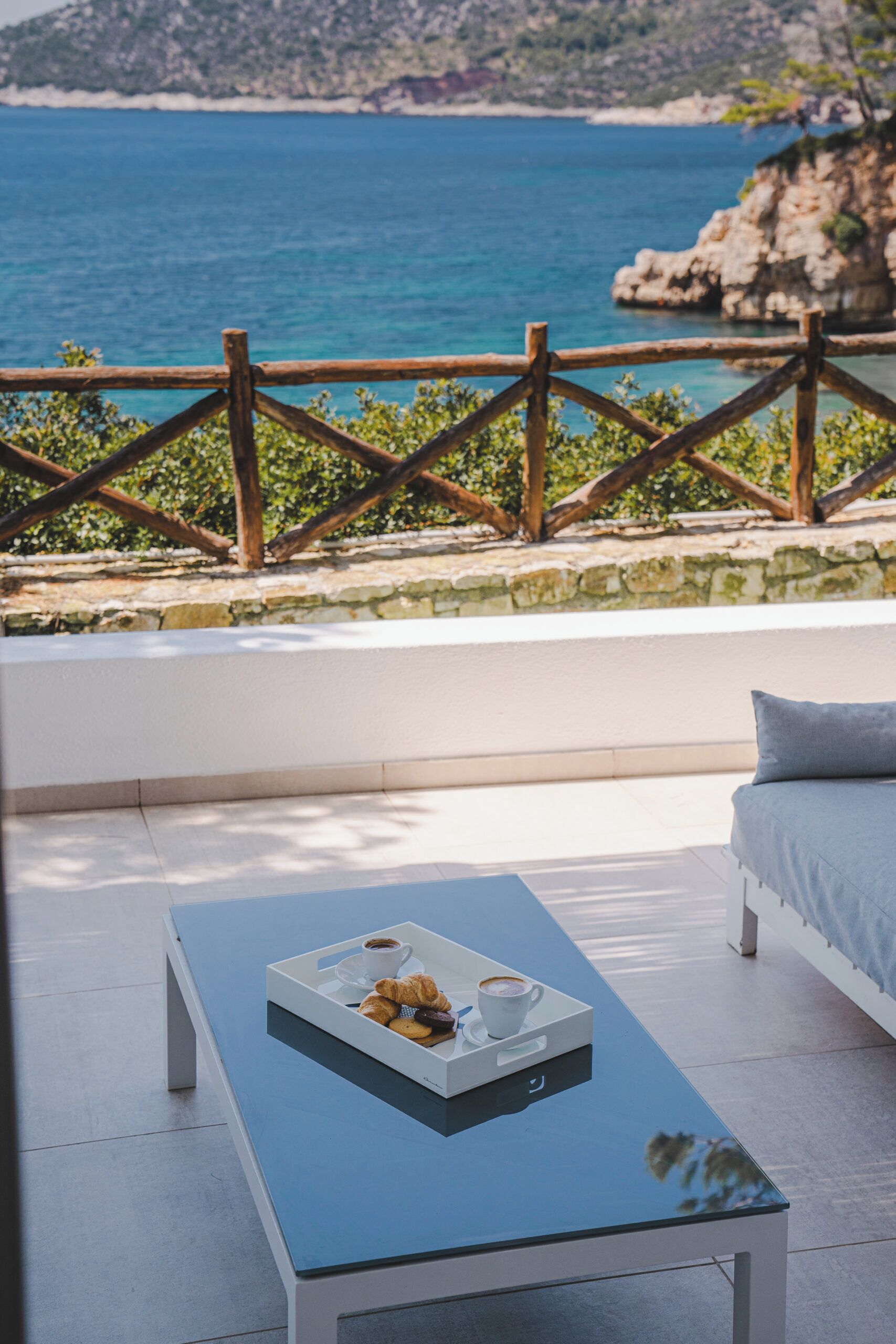 A modern outdoor seating area with a glass table, a white sofa, and a view of the sea.