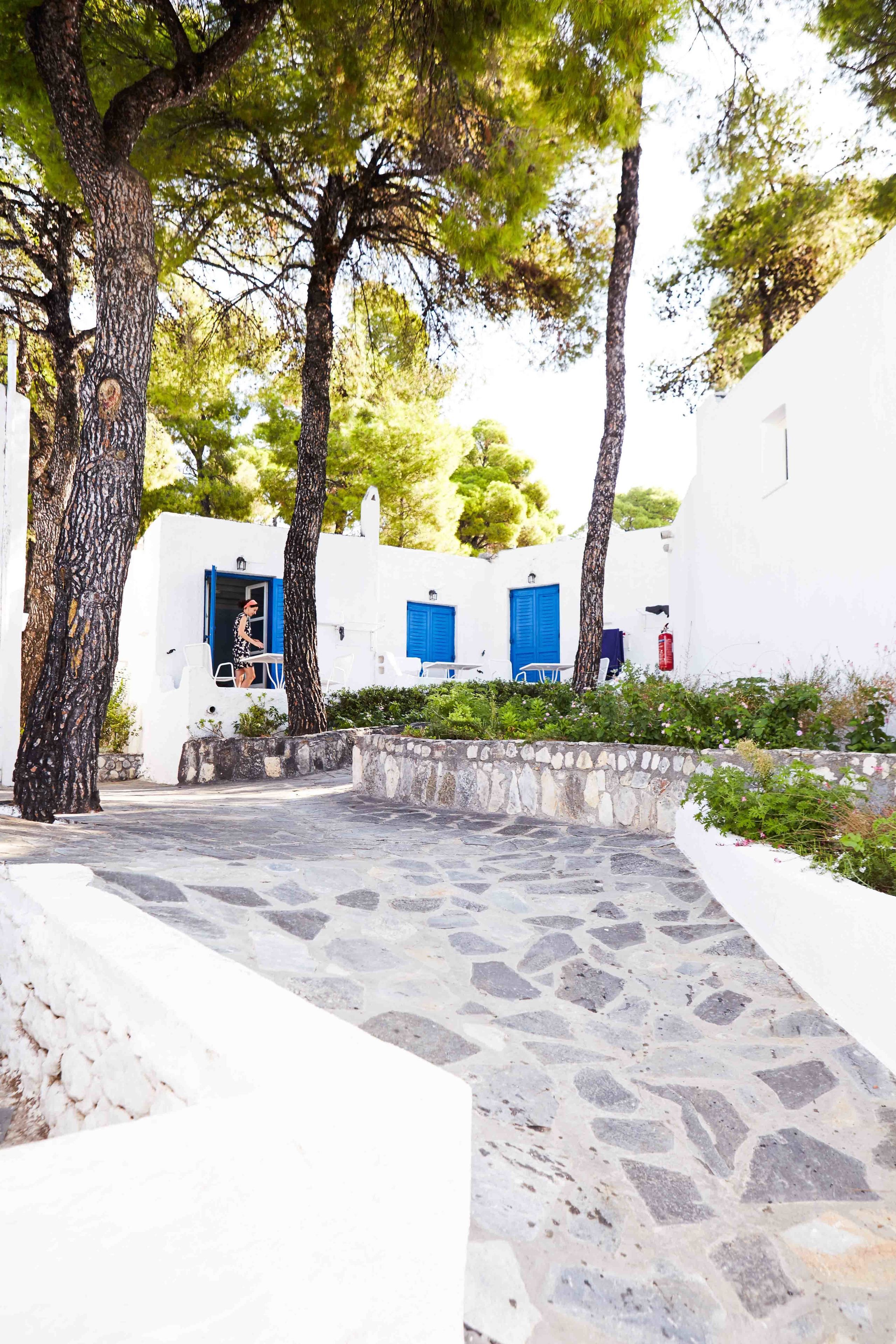 A narrow stone-paved pathway leading to white buildings with blue doors and windows, surrounded by trees and greenery.