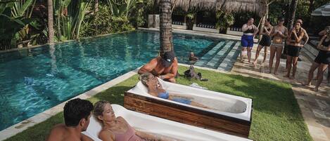 Zona de piscina con sillones reclinables, una bañera de hidromasaje y un grupo de personas junto a la piscina.