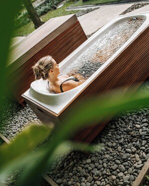 A person relaxing in a hot tub outdoors surrounded by rocks and wooden structures. - Hotel Komune and Beach Club Bali (Keramas)