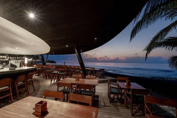 Une terrasse en bois, avec des tables et des chaises, donnant sur l’océan au coucher du soleil.