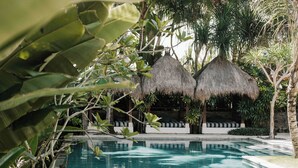 A swimming pool with thatched-roof structures in the background, surrounded by lush greenery.