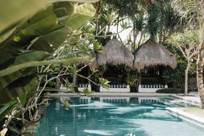 Una piscina con estructuras de techo de paja al fondo, rodeada de exuberante vegetación.