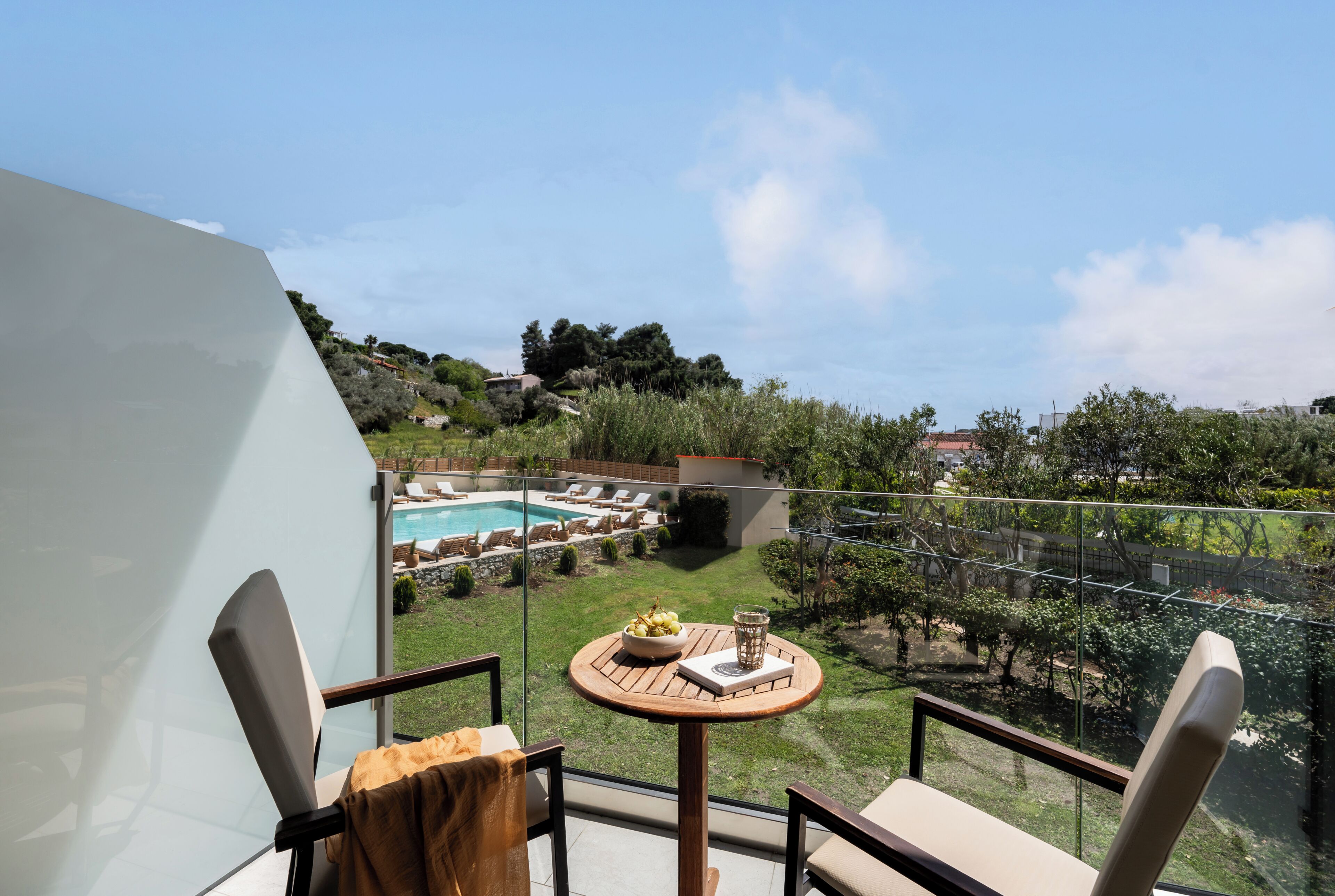 A balcony with a glass railing, a round wooden table with a bowl of fruit and a glass of water, and a view of a swimming pool and garden.