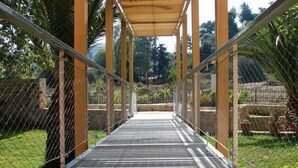 Eine Fußgängerbrücke mit Metallgeländern und einem hölzernen Vordach, umgeben von Grünflächen und einem Maschendrahtzaun.
