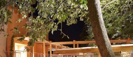 Un área de comedor al aire libre con mesas y sillas, rodeada de árboles y una pared de piedra.