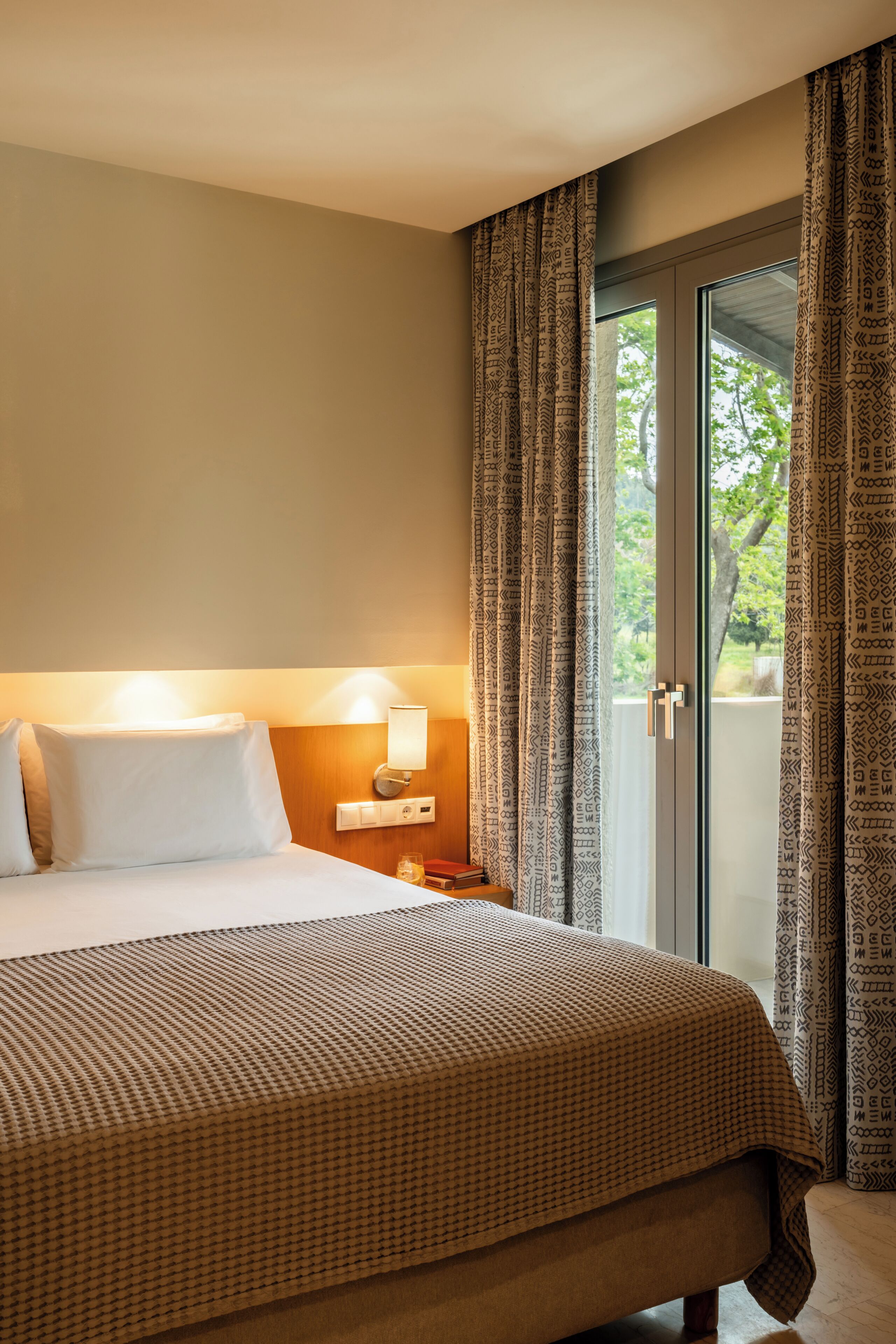 Ein Hotelzimmer mit einem Bett, Nachttischlampen, einem Beistelltisch und Blick auf Grünanlagen durch das Fenster.