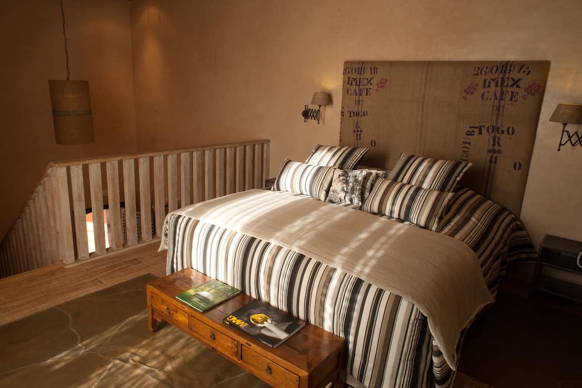 Une chambre avec un grand lit, une literie à rayures, un banc en bois avec des livres, une lampe suspendue et un mur orné d’une vieille enseigne.