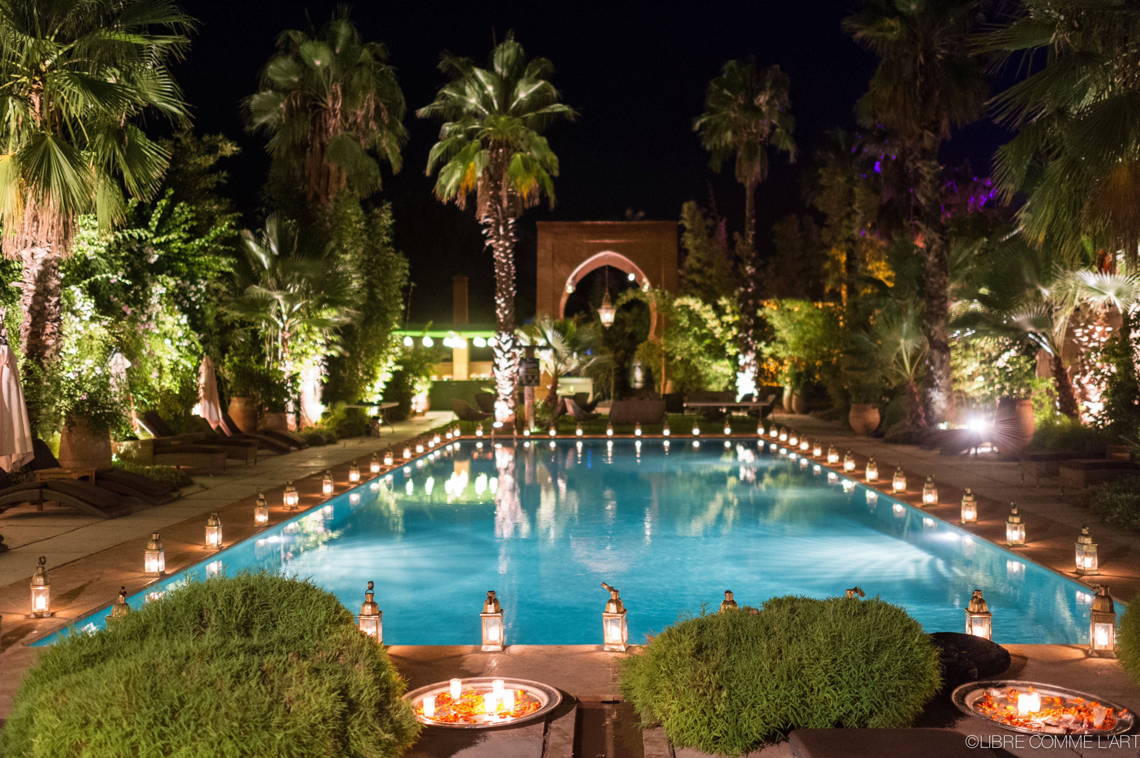 Zona de piscina nocturna con piscina cristalina, rodeada de palmeras e iluminada por faroles.