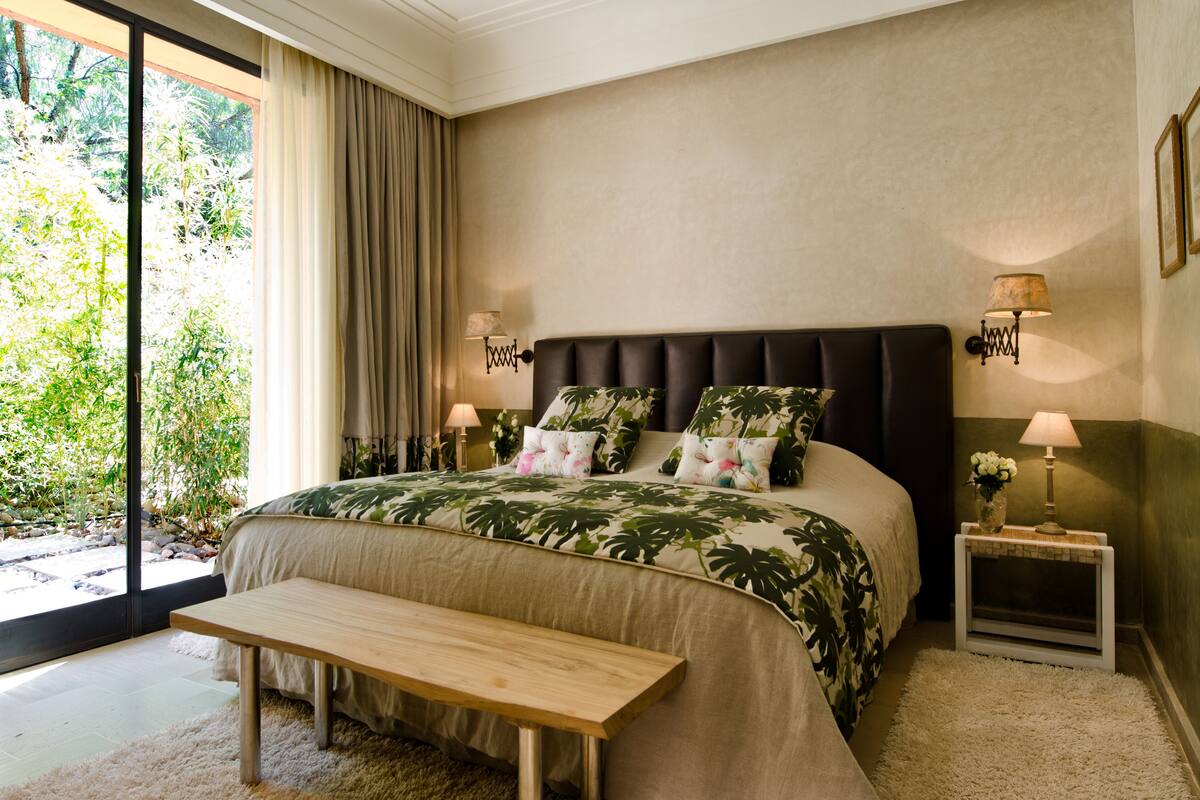 Une chambre avec un grand lit, un banc, deux tables de chevet avec des lampes et une vue sur la verdure grâce à une porte coulissante.