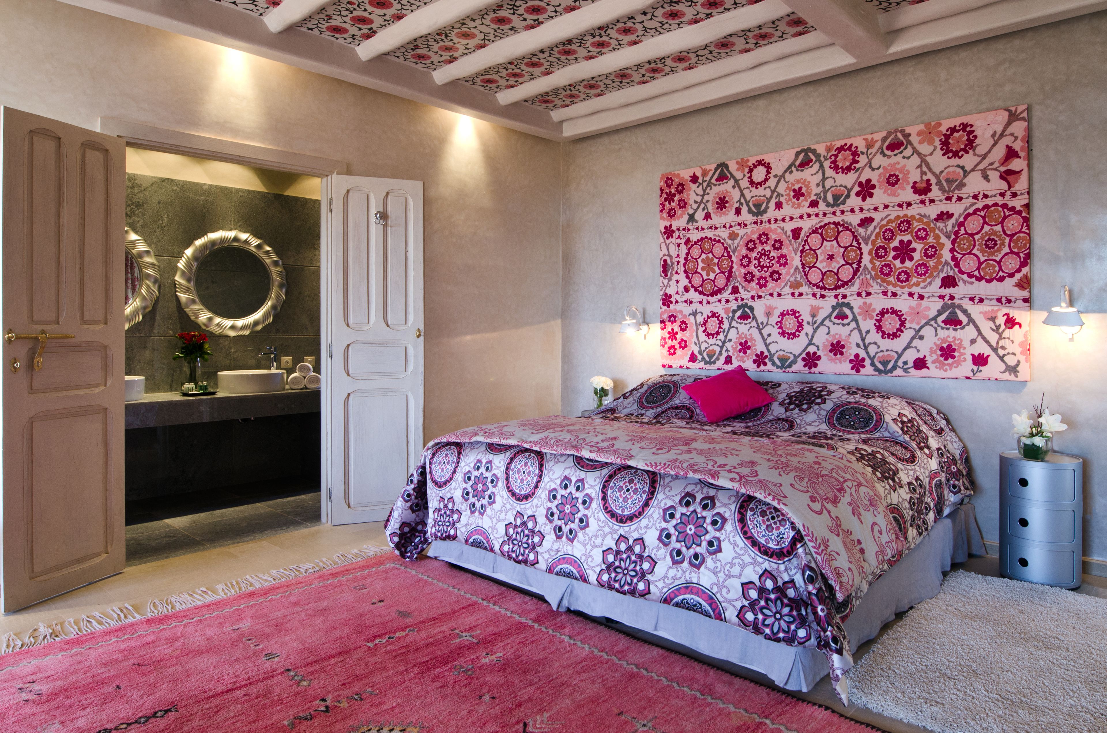 Un dormitorio con cama, mesita de noche, tapiz en la pared, espejo y puerta que da al baño.