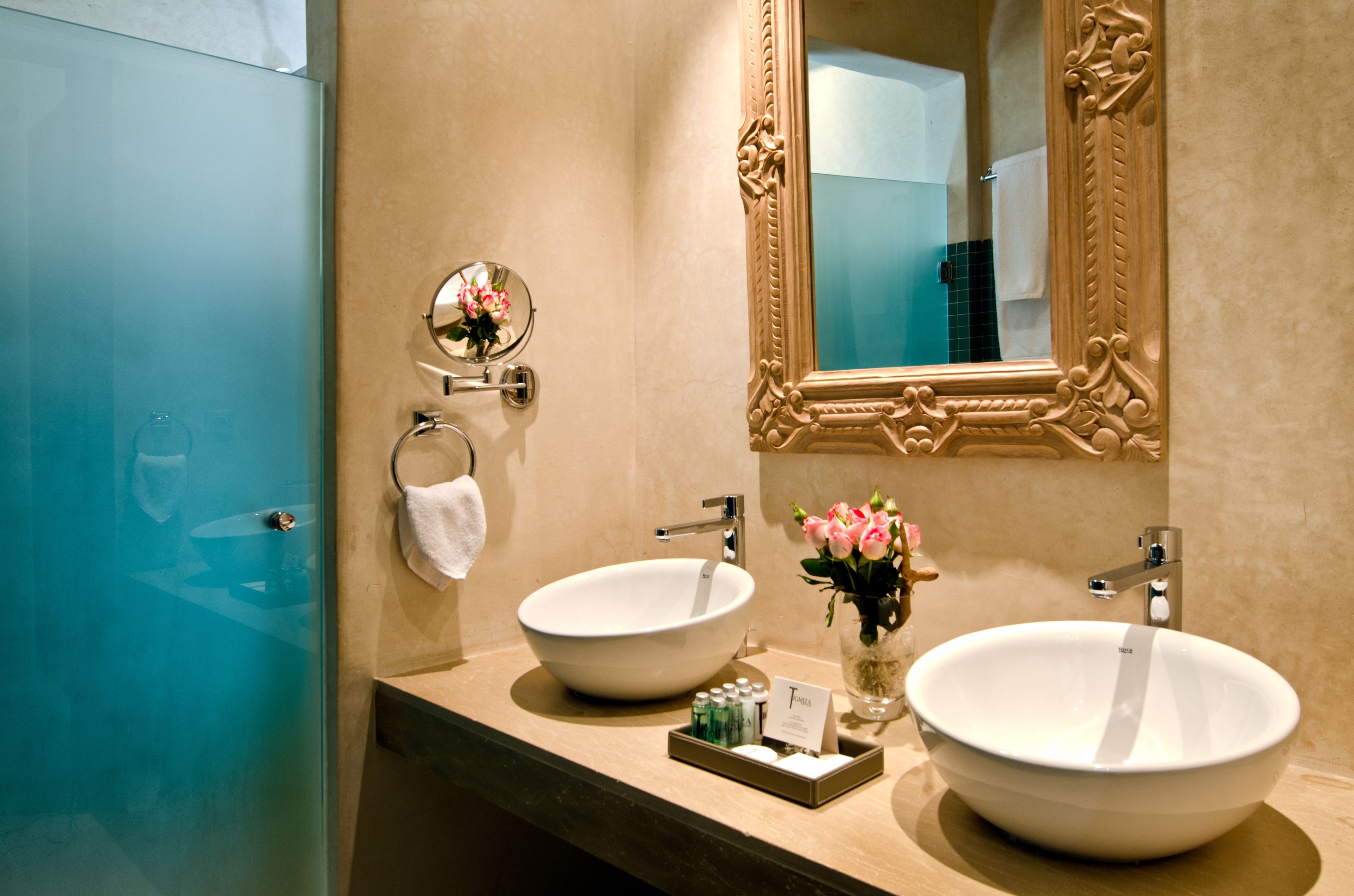 Une salle de bain avec deux grands lavabos blancs, un miroir doté d’un cadre orné et un petit vase avec des fleurs.