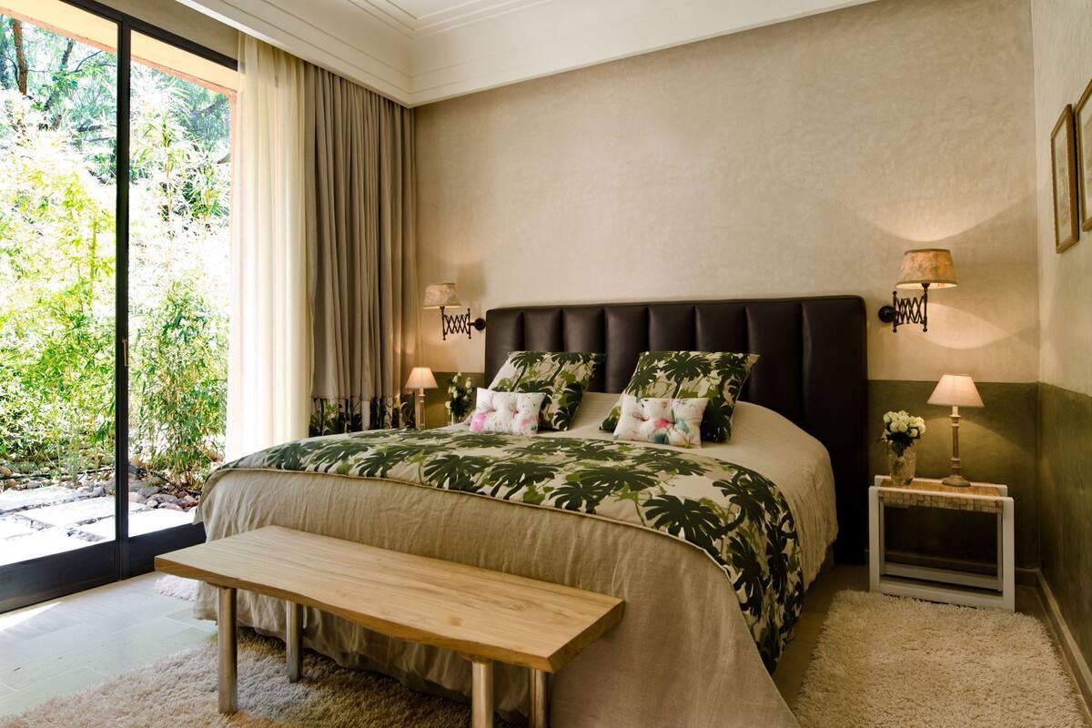 Une chambre avec un grand lit, un banc, deux tables de chevet avec des lampes et une vue sur la verdure.
