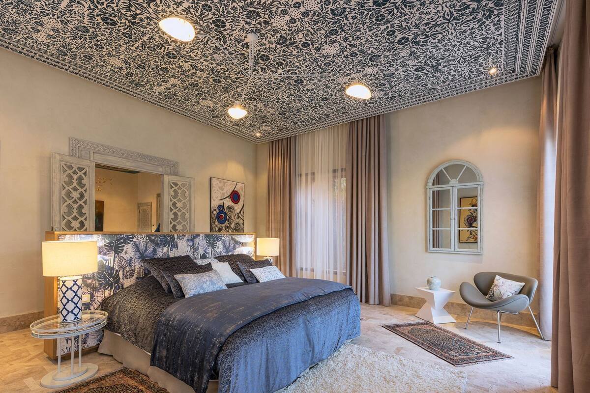 Une chambre à coucher avec un plafond décoré, un grand lit avec de nombreux oreillers, une table de chevet avec une lampe, une chaise et une fenêtre avec des rideaux.