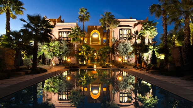 Un hotel lujoso con piscina reflectante, palmeras y una entrada bien iluminada.