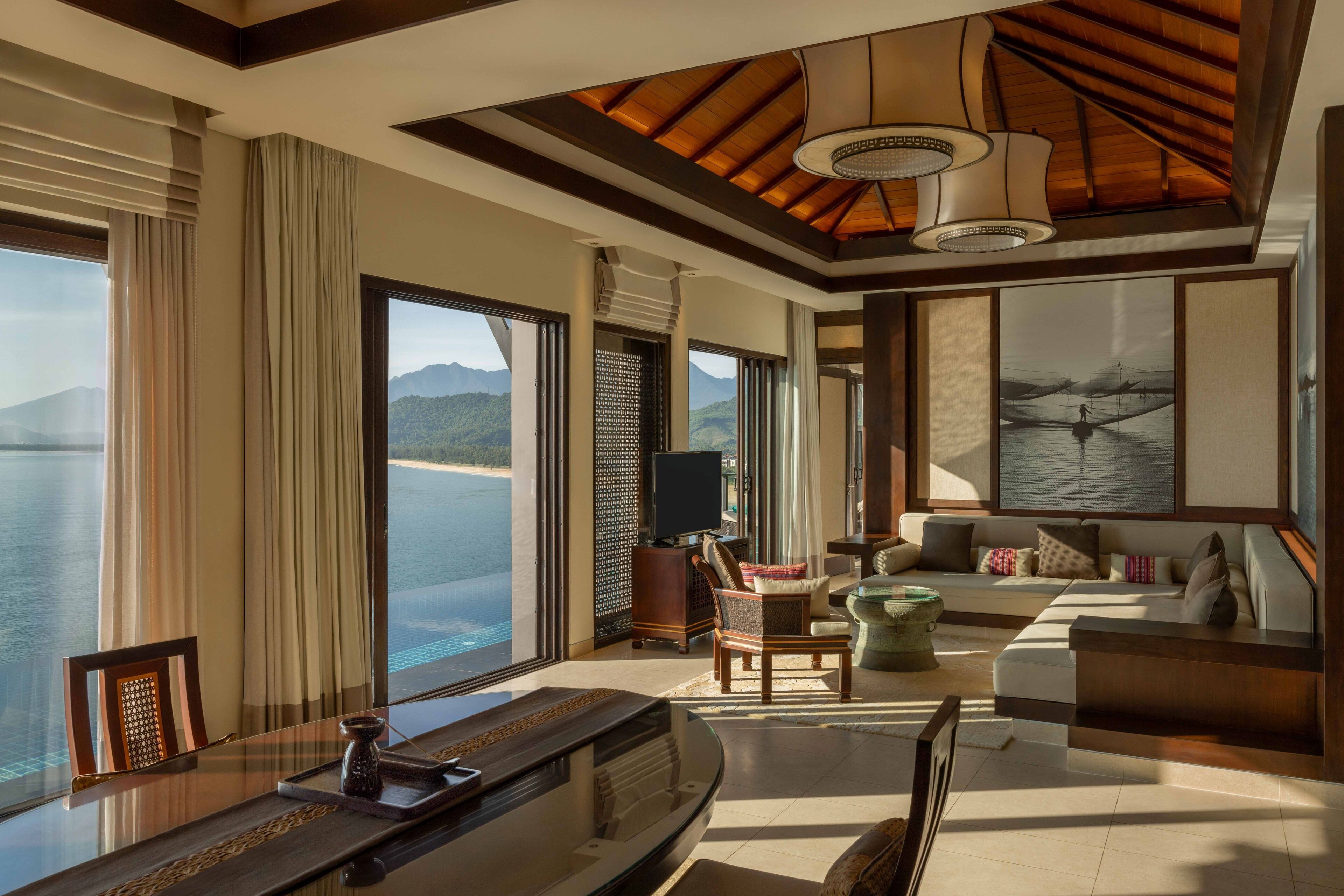 A spacious room with a large window offering a view of the sea and mountains, a comfortable seating area, and a television on a wooden stand.