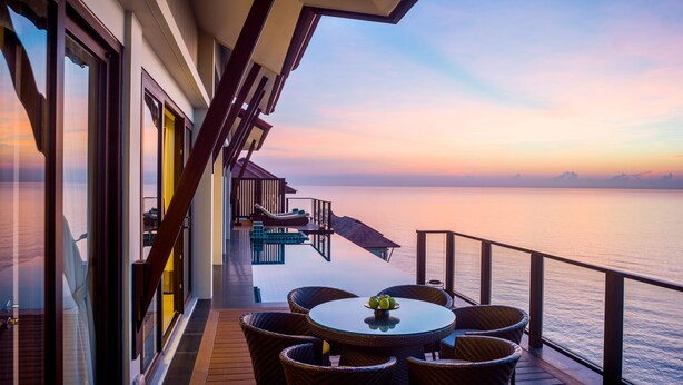 Un balcón con sillas de mimbre y una mesa, con vista al mar tranquilo al atardecer.
