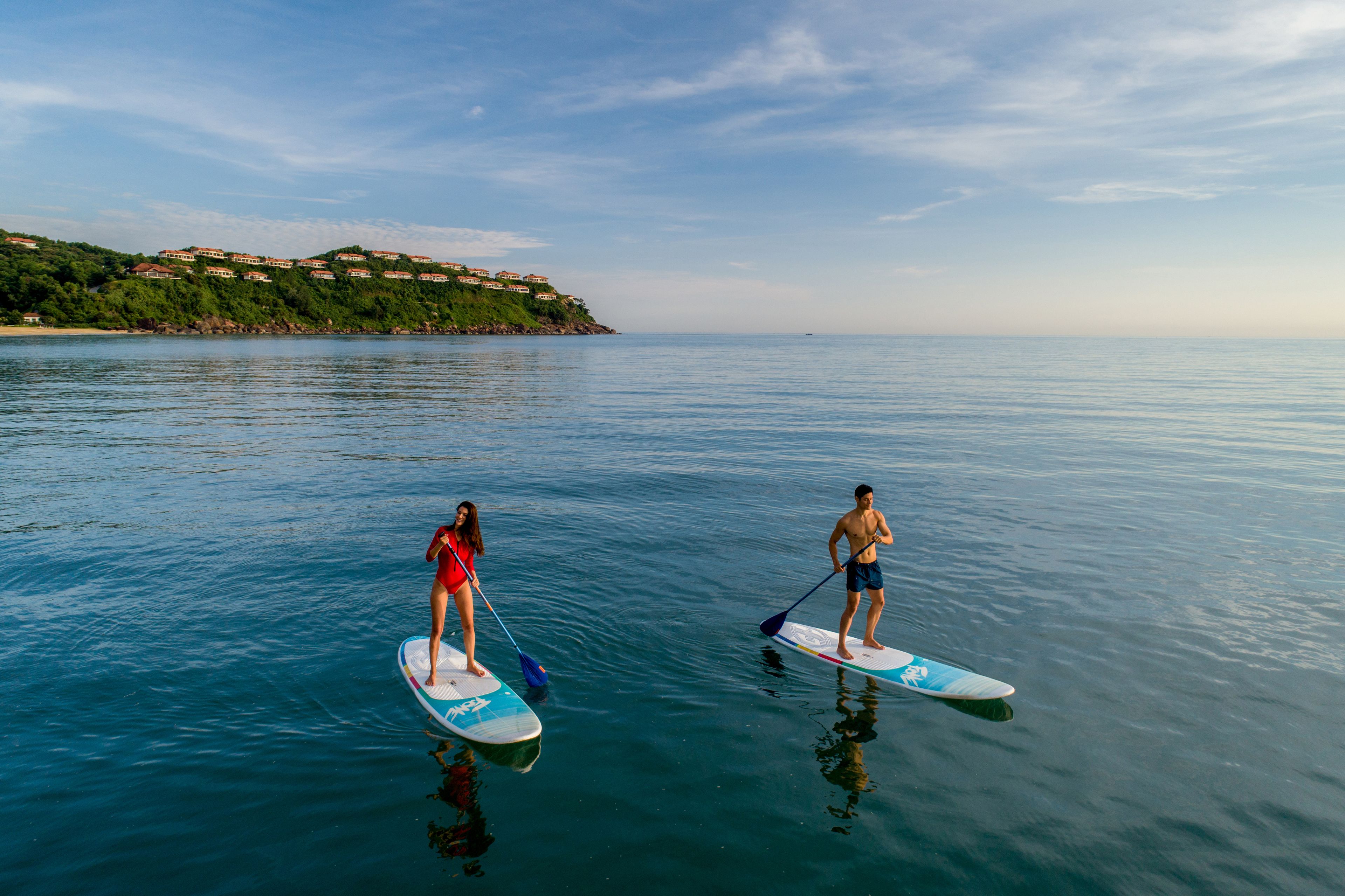 Two people paddleboarding on a calm sea with a coastal landscape in the background.