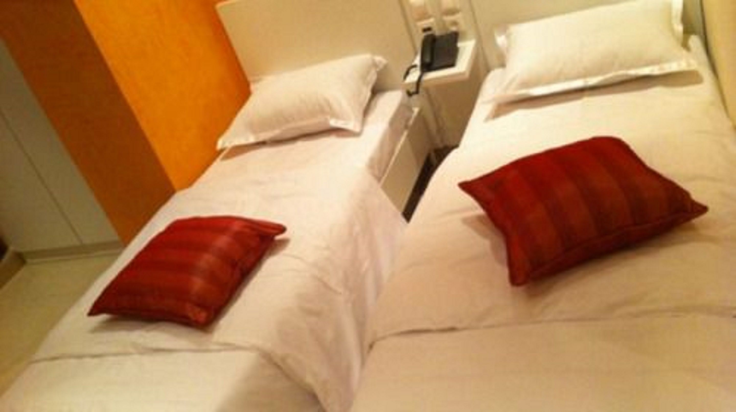 Two twin beds with white linens and red pillows, a bedside table with a lamp, and a wooden wall.