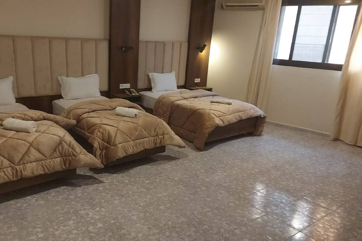 A hotel room with three beds, each with a beige quilted duvet and white pillows. The room has a tiled floor and a wooden headboard.