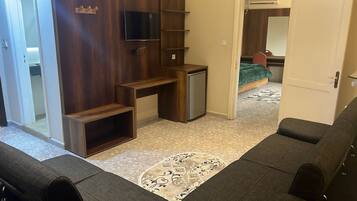 A compact living space with a kitchenette, a TV mounted on the wall, a bed with a green blanket, and a patterned rug.