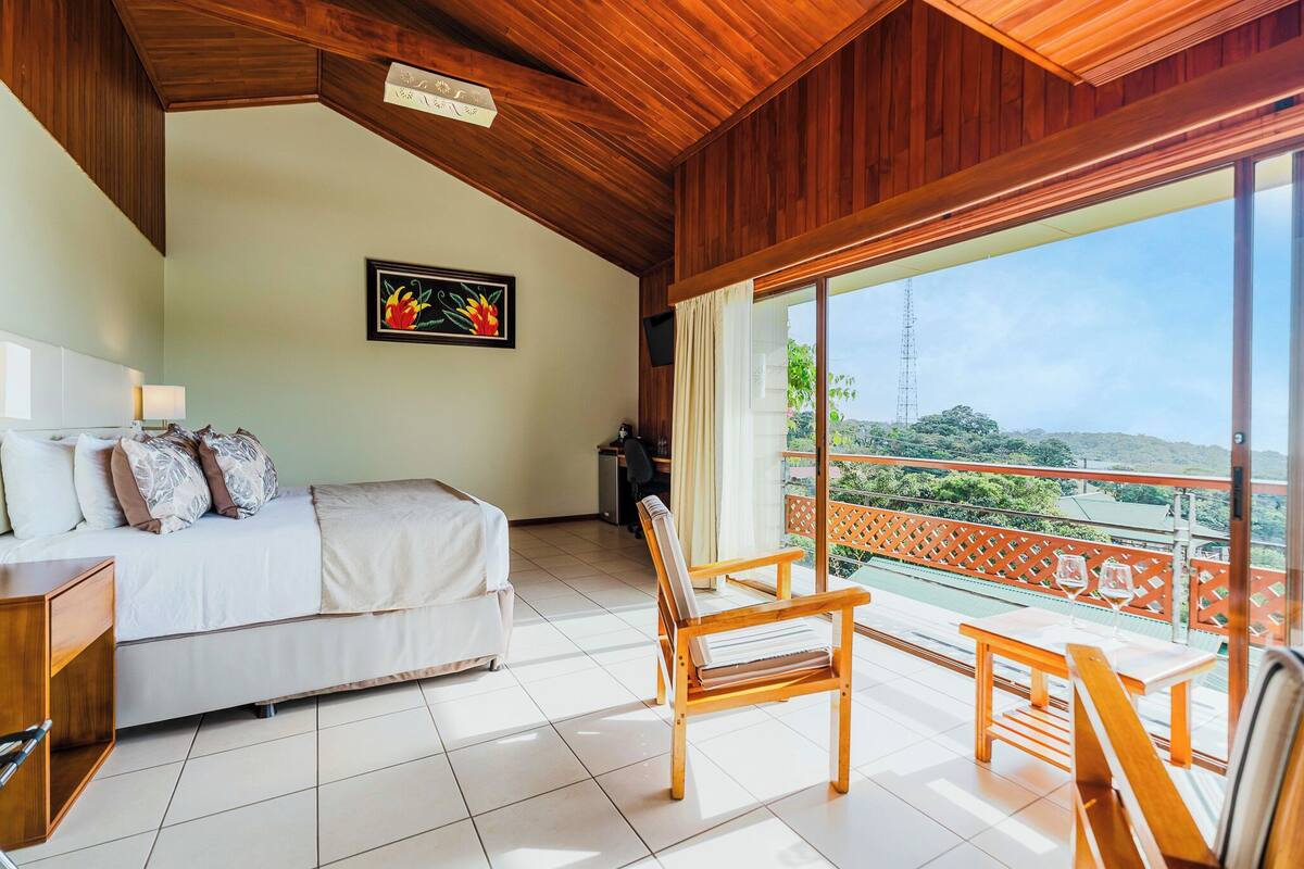 A bedroom with a bed, a desk, and a chair. There is a balcony with a view of the landscape.