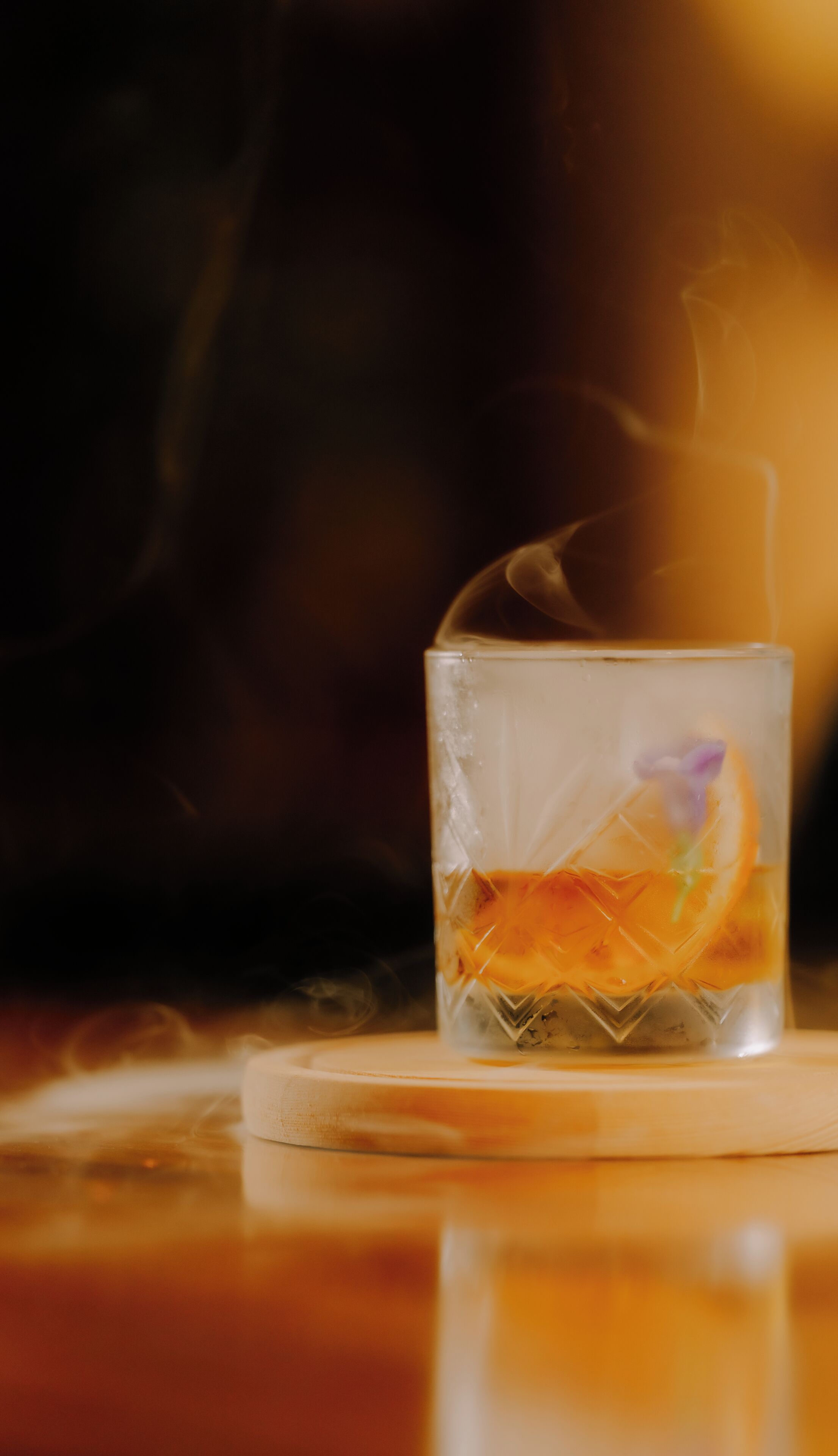 A glass with a slice of orange and a purple flower, steam rising from it, placed on a coaster.
