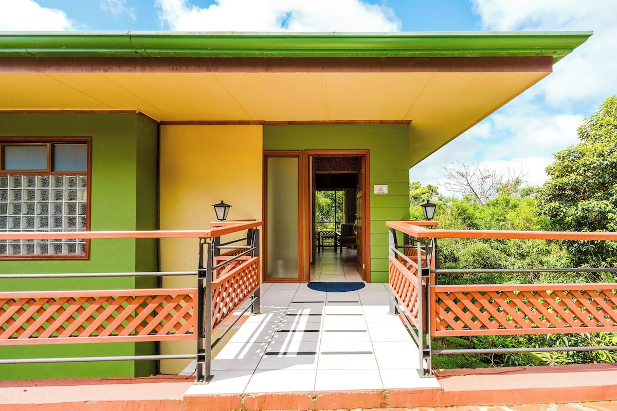 A building with a green and yellow facade, a covered entrance with a tiled floor, and a view of greenery in the background.
