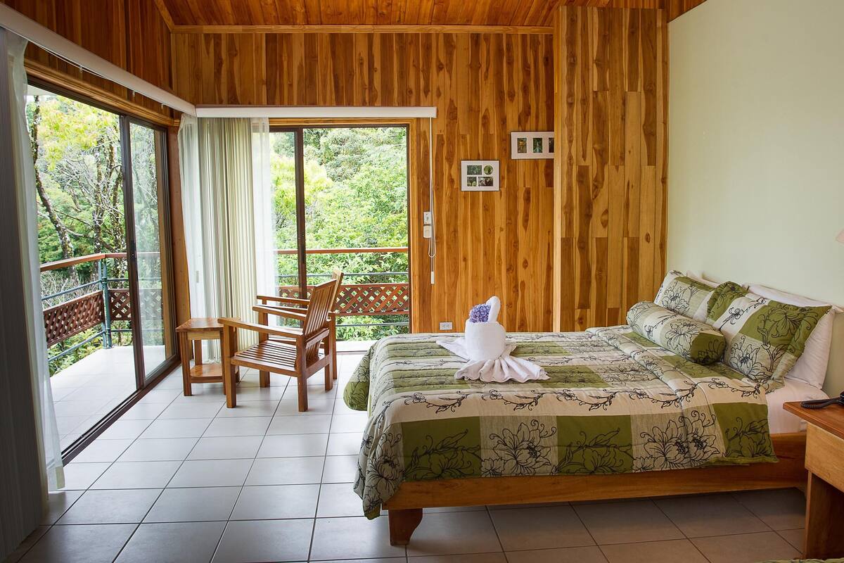 A wooden-paneled room with a bed, a chair, and a balcony with a view of trees.
