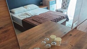 A hotel room with a wooden table, a bed with a blue headboard, a chair, and a window with curtains. - Kleopatra Bebek Hotel (Alanya)