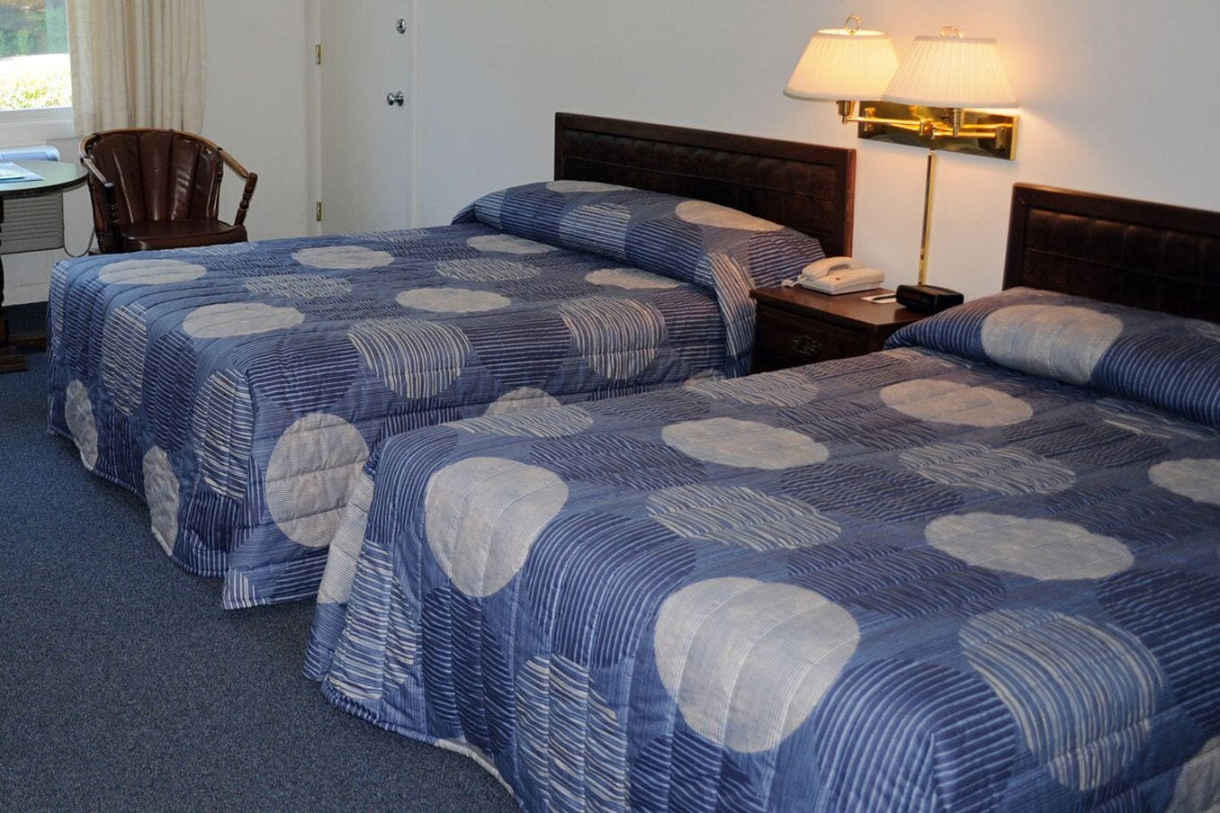 A hotel room with two beds, a desk, a chair, and a lamp.