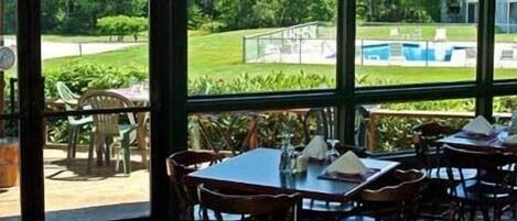 Espaço de refeições interno com mesas e cadeiras, grandes janelas com vista para uma piscina e vegetação.