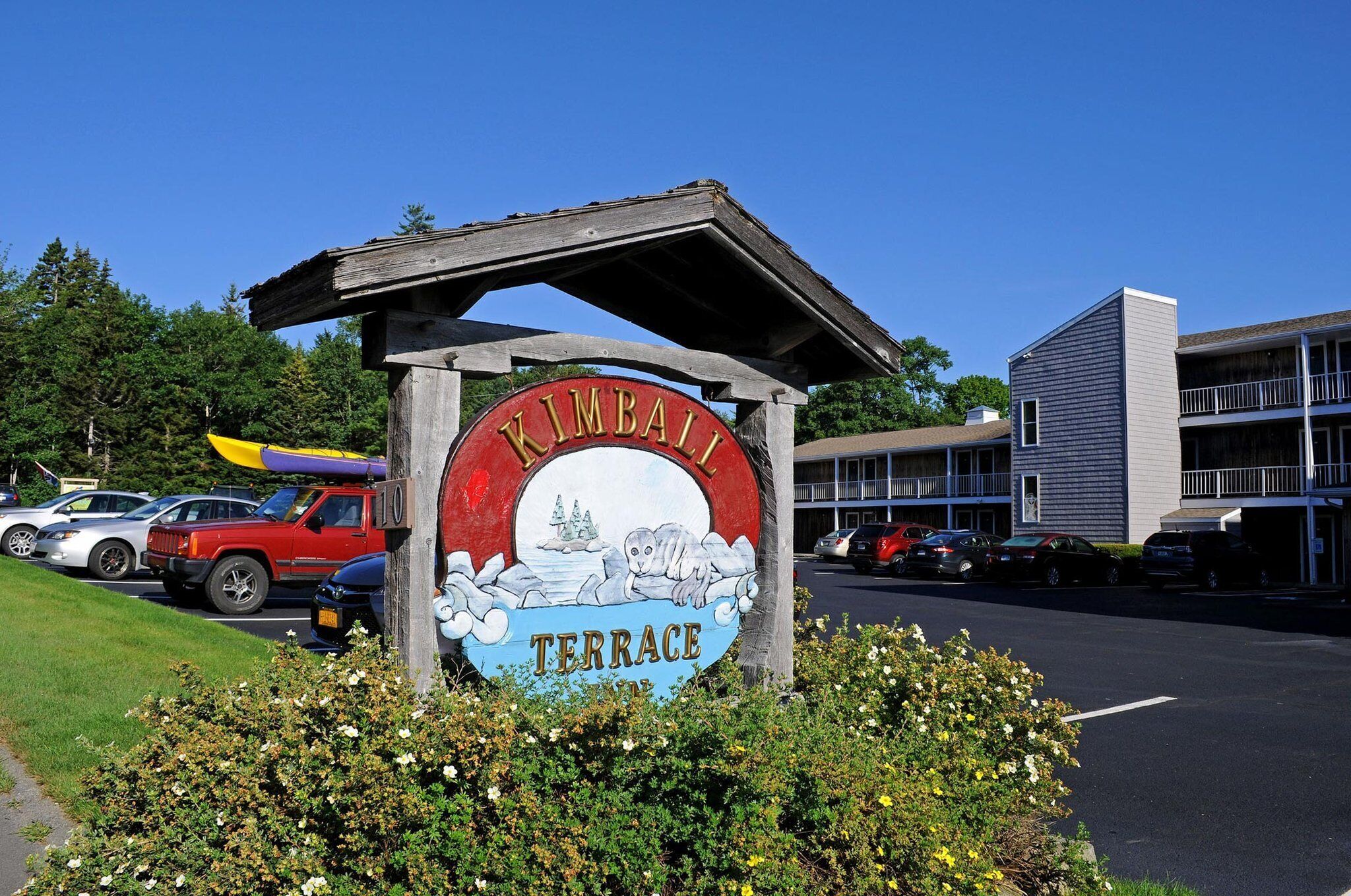 A sign for Kimball Terrace with a kayak and seal illustration, surrounded by parked cars and a building in the background.