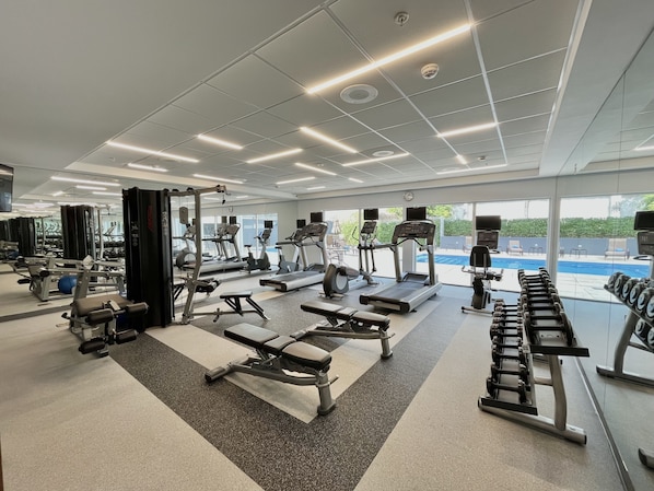 A modern gym with various exercise equipment, large windows, and a view of an outdoor swimming pool.
