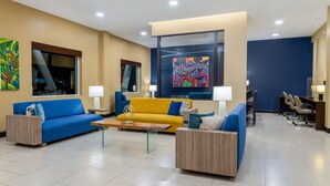 A modern hotel lobby with a yellow sofa, blue armchairs, and a wooden coffee table. There are two paintings on the walls and a large window. - Courtyard by Marriott San Jose Airport Alajuela (Rio Segundo)